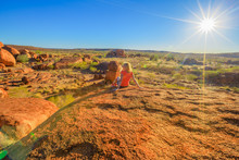 Tourist Woman On The Top Of Karlu Karlu Devils Marbles Conservation Reserve Looking Panoramic Views Of Granite Boulders Rock Formations At Sunset With Sunrays Of Outback. Northern Territory, Australia