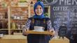 © PRPicturesProduction - attractive muslim female worker welcoming customer to modern coffeehouse. young malay girl in blue hijab holding tray with cup of coffee on it sending to camera smiling. service cafe concept