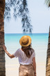 © allenkayaa - A young woman under a palm tree on the shore of the Gulf of Thailand. Woman in a hat looks at the sea