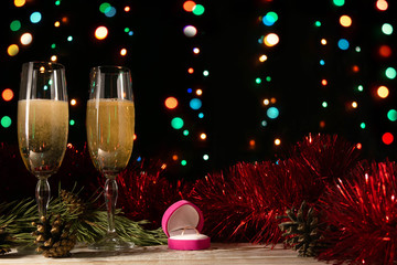 Proposal to get married on New Year's or Christmas night. Dinner of a couple with two glasses of champagne and a wedding ring. Tinsel and spruce branch on a black background with blurred lights.