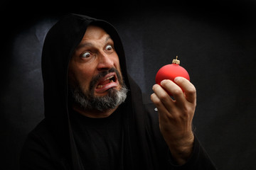 Wall Mural - Portrait of a bald man with a beard in a black hood witha red christmas ball on a dirty gray background. Young Santa concept.