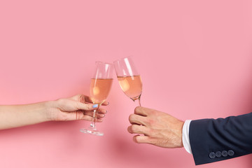 Cropped hands holding glass of champagne isolated over pink background. Unrecognizable couple of man in tuxedo drink glass of champagne with female