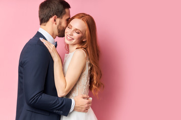 Wall Mural - Caucasian man in suit and woman in white wedding dress with long hair lovely hug each other and smile, after wedding ceremony posing isolated over pink background.