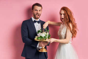 Wall Mural - Portrait of caucasian couple, bearded man in tuxedo surprised, woman with red hair want sweet cake. isolated over pink background