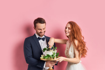 Wall Mural - Happy couple holding sweet cake isolated over pink background. Woman with auburn hair and bearded man in wedding day. Celebration, party, love concept