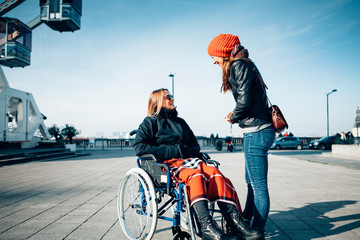 two smiling women talking and spending time together outdoor in the city - sitting on a wheelchair and living a disability with positive attitude concept