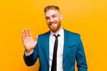 Young Red Head Businessman Smiling Happily And Cheerfully, Waving Hand, Welcoming And Greeting You, Or Saying Goodbye Against Orange Background