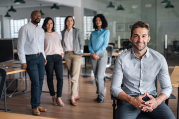 Smiling businessman sitting in an office with coworkers behind him