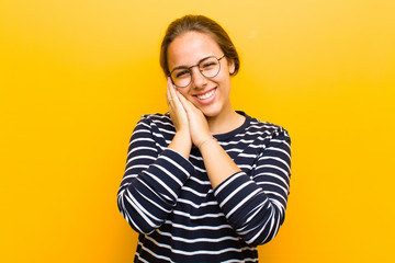 Wall Mural - young pretty woman feeling in love and looking cute, adorable and happy, smiling romantically with hands next to face against orange background