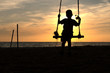© Mitch Saint - Silhoutte image of young boy having fun on the swing with water bottle in hand, kid playing wooden swing on the beach in sunset.