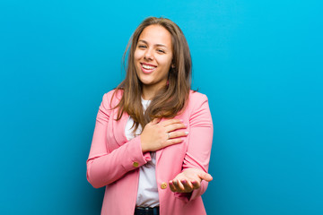 Wall Mural - young woman feeling happy and in love, smiling with one hand next to heart and the other stretched up front against blue background