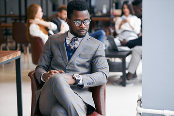 Portrait of handsome black man in eyeglasses wearing grey formal suit, elegant and serious businessman while his business partners have conversation behind him