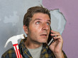 © TheVisualsYouNeed - funny portrait of messy and frustrated man calling domestic insurance on mobile phone for repair mess he did with a hammer cracking the wall needing professional help