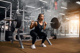 Beautiful young powerlifter squatting in modern fitness studio using heavy barbell, looking aside expressing self assurance, professional sport concept, white smoke in the air