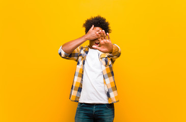Wall Mural - young black man covering face with hand and putting other hand up front to stop camera, refusing photos or pictures against orange wall
