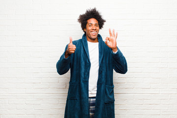 Wall Mural - young black man wearing pajamas with gown feeling happy, amazed, satisfied and surprised, showing okay and thumbs up gestures, smiling against brick wall