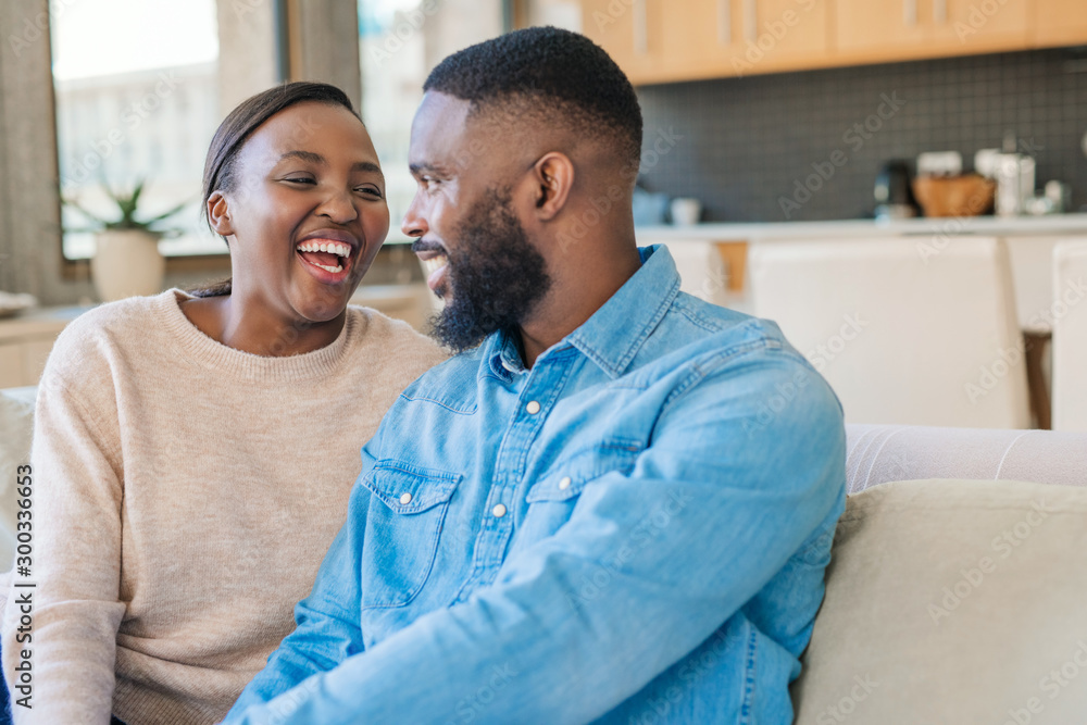 Young African American couple sitting on their sofa and laughing - obrazy, fototapety, plakaty 