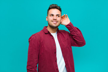 Wall Mural - young hispanic man smiling, looking curiously to the side, trying to listen to gossip or overhearing a secret against blue background