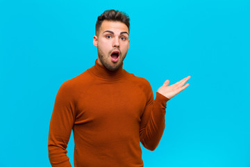 Wall Mural - young hispanic man looking surprised and shocked, with jaw dropped holding an object with an open hand on the side against blue background