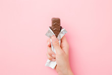 Young Woman Hand Holding Chocolate Bar On Pastel Pink Table. Opened Pack. Sweet Snack. Close Up.
