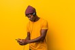 © Asier - Young african man standing against a yellow background wearing a hat listening to music with a phone