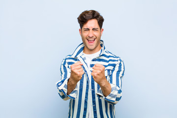 Wall Mural - young handsome man shouting triumphantly, laughing and feeling happy and excited while celebrating success against blue background