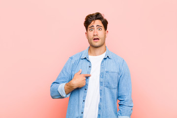 Wall Mural - young handsome man looking shocked and surprised with mouth wide open, pointing to self against pink background