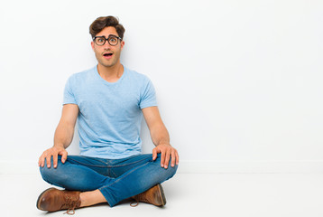 Wall Mural - young handsome man looking happy and pleasantly surprised, excited with a fascinated and shocked expression sitting on the floor