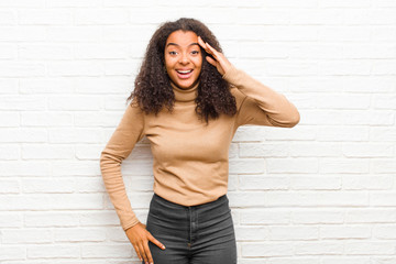 Wall Mural - young black woman looking happy, astonished and surprised, smiling and realizing amazing and incredible good news against brick wall