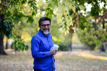 Wall Mural - Happy senior smiling businessman going to the work with coffee cup and outdoors, successful employer  on break outside at sunny day near his office in beautiful park