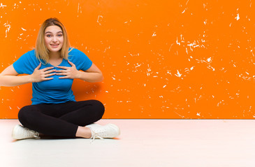 Wall Mural - young blonde woman looking happy, surprised, proud and excited, pointing to self sitting on the floor