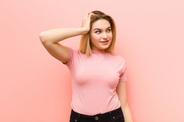 Wall Mural - young pretty blonde woman smiling cheerfully and casually, taking hand to head with a positive, happy and confident look against pink flat wall