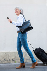 Wall Mural - Full length older woman walking with mobile phone and luggage on street