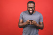 Portrait of handsome excited cheerful joyful delightful african american guy wearing casual sending and getting messages to his lover isolated against red background. Using phone.
