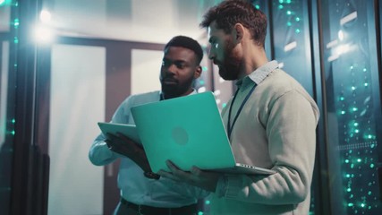 Two IT technicians cooperating collecting network data from rack server cabinets using laptop and tablet inspecting data center computer security.