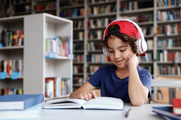 Wall Mural - Cute little boy with headphones reading books at table in library