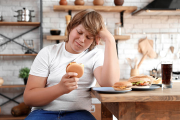 Wall Mural - Emotional overweight boy at table with fast food in kitchen