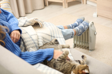 Sticker - Young woman with cat warming feet on electric heater at home, closeup