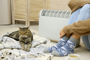 Canvas Print - Young woman and cute tabby cat near electric heater at home, closeup