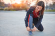 © astrosystem - Young modern woman tying running shoes in urban park.