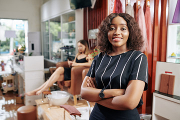 portrait of pretty young manicurist smiling at camera, her client waiting for pedicure in armchair