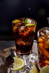 selective focus of cocktails cuba libre in glasses with straws, ice cubes and limes on black background