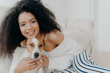 Wall Mural - Image of attractive dark skinned woman wears makeup, has bushy curly hair, smiles pleasantly, cuddles dog, dressed in fashionable clothes, enjoys sweet moment with puppy, being at home together