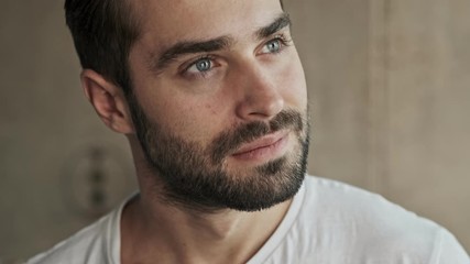 Poster - Close up view of thougthful handsome young brunet man smiling and touching his beard while looking to the side at home