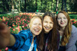 © exebiche - Happy young girls have fun laughing and taking selfies on a smartphone in the park on the background of green foliage and flowers. Blonde, brunette and girl with red hair, female friendship concept.