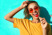Portrait Of A Young Stylish Woman Smiling At Camera, Licking Big Red Lollipop Against Blue Background.