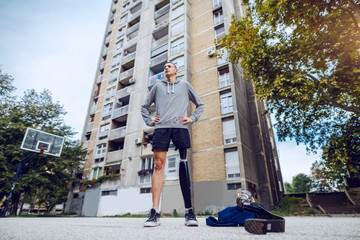 Wall Mural - Low angle view of handsome caucasian sporty young handicapped man with artificial leg and in sportswear standing with hands on hips on basketball court.