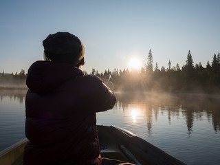 Fisherman man in rowing boat at lake Sjabatjakjaure in Beautiful sunny morning haze mist in Sweden Lapland nature. Mountains, birch trees, spruce forest. Blu sky and backlight