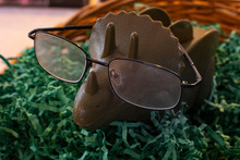 A Green Plastic Triceratops Wearing An Old Pair Of Black Rimmed Glasses In A Basket Filled With Green Paper Filler.
