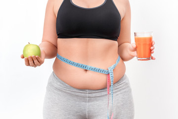 Wall Mural - Body Care. Chubby girl with bare belly and tape measure on waist standing isolated on white holding apple andsmoothie close-up